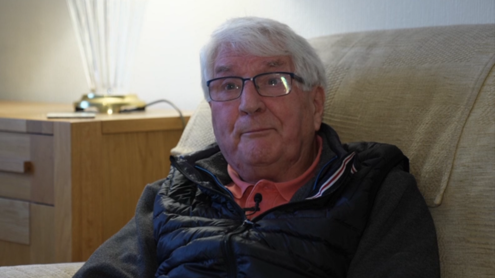 A man who appears to be in his sixties or early seventies, Stuart Couzens. He has wispy white hair which falls across his forehead. He wears thin framed, black glasses. He has a kind looking face. He wears a black gilet, dark grey jumper, and coral polo neck shirt.