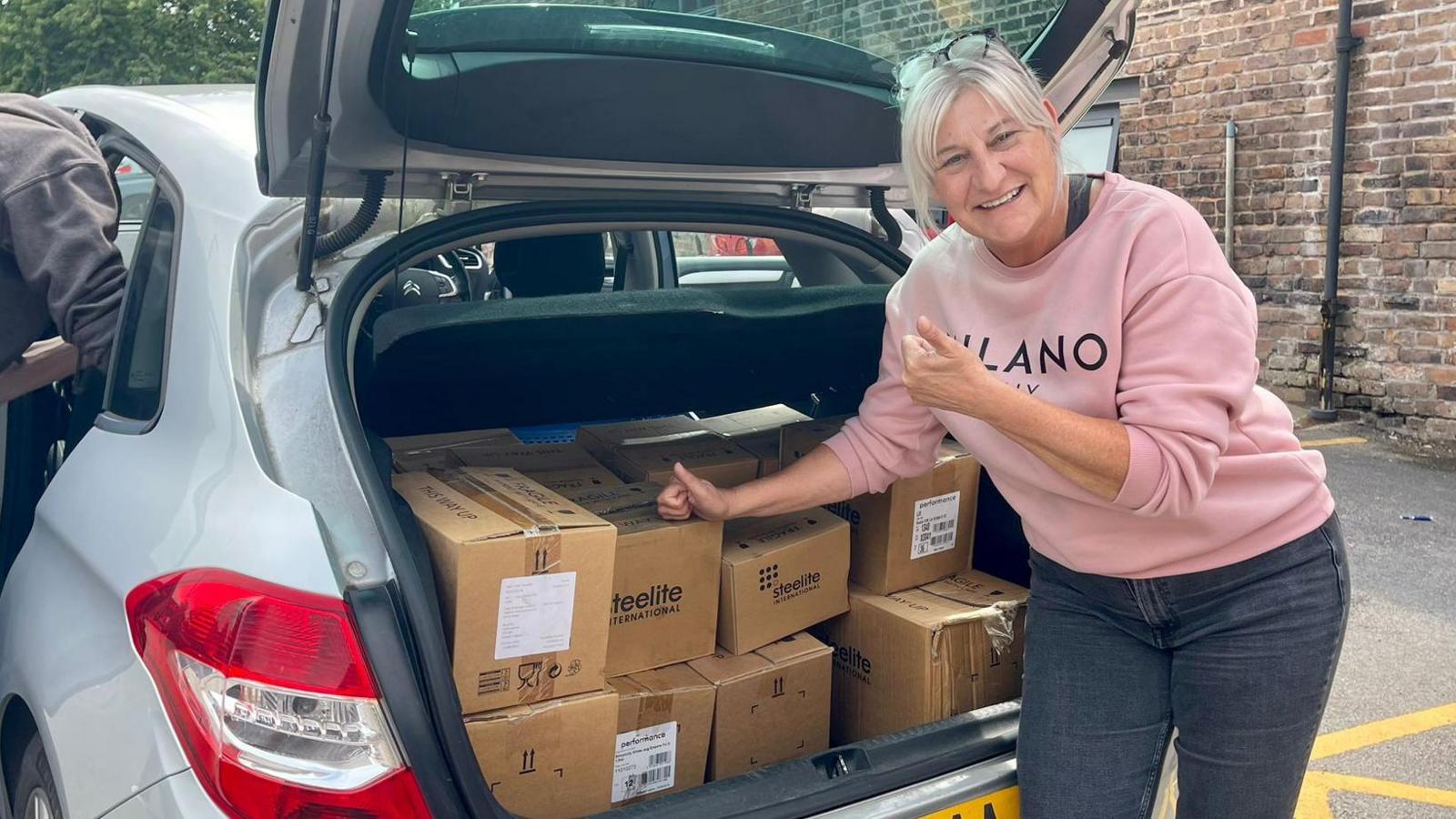 A woman in a pink jumper has her thumbs up, standing beside the boot of a silver car, filled with cardboard boxes printed with the Steelite logo. The woman has short white hair tied back, blue jeans and glasses perched on her head.