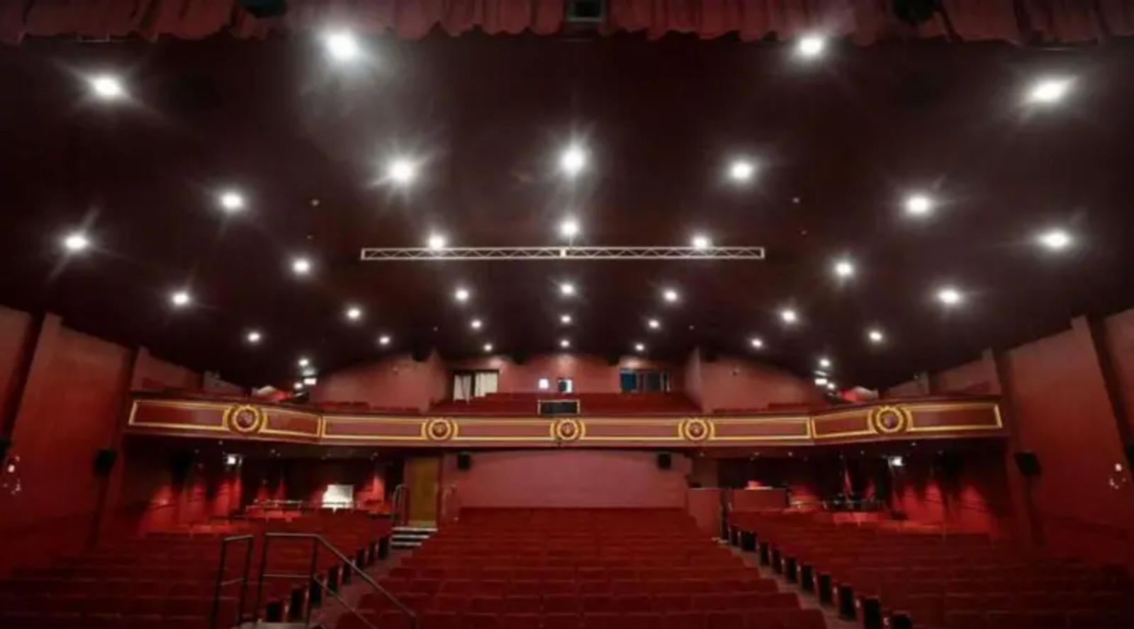 The auditorium of the Empire in Consett pictured from the stage and showing several rows of red seating and a red-painted balcony front which features a gold pattern. There are multiple lights in the ceiling.