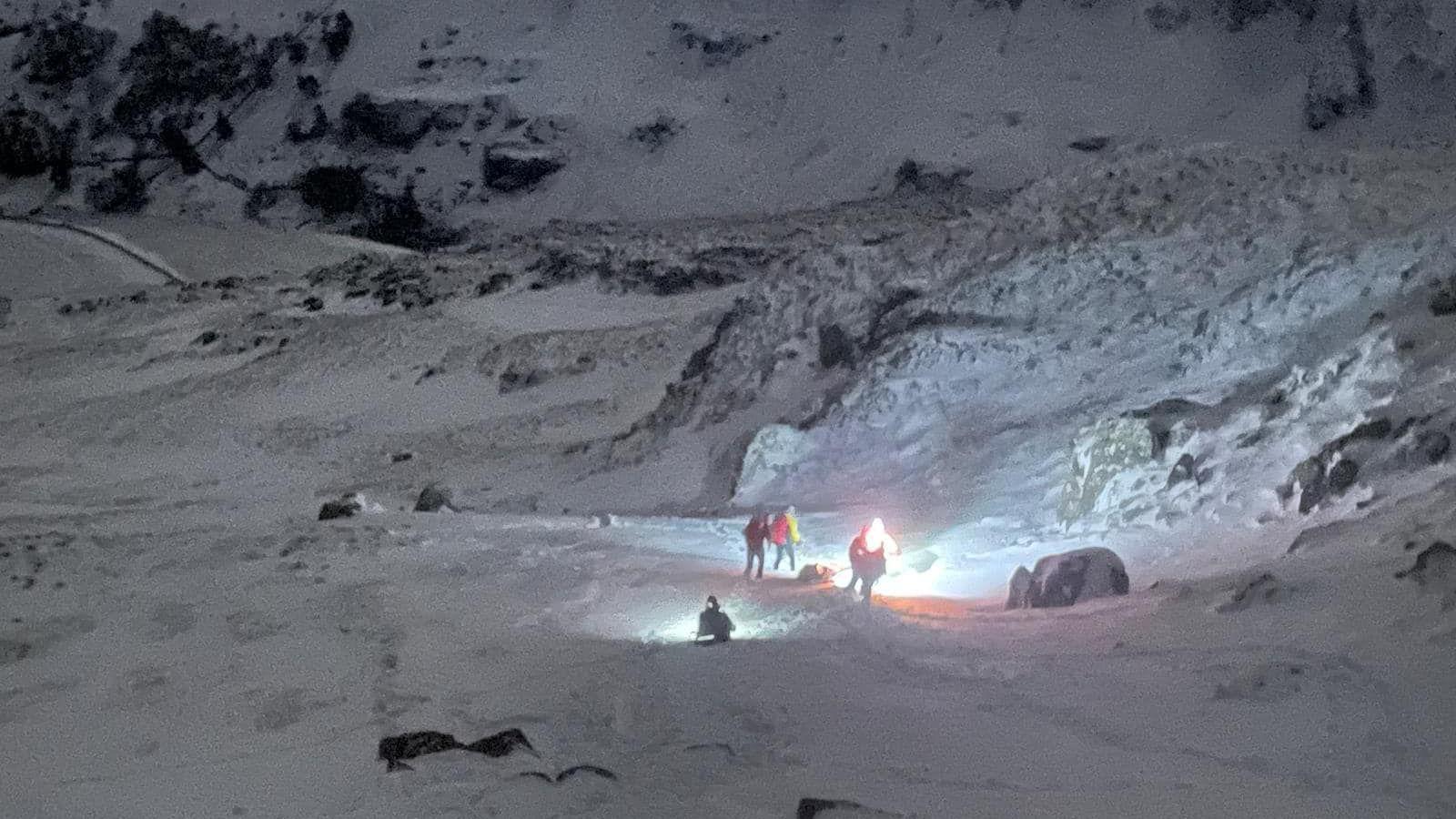 Snowy mountain in the dark. In the foreground are people in orange jackets with torchlights looking at the snow. 