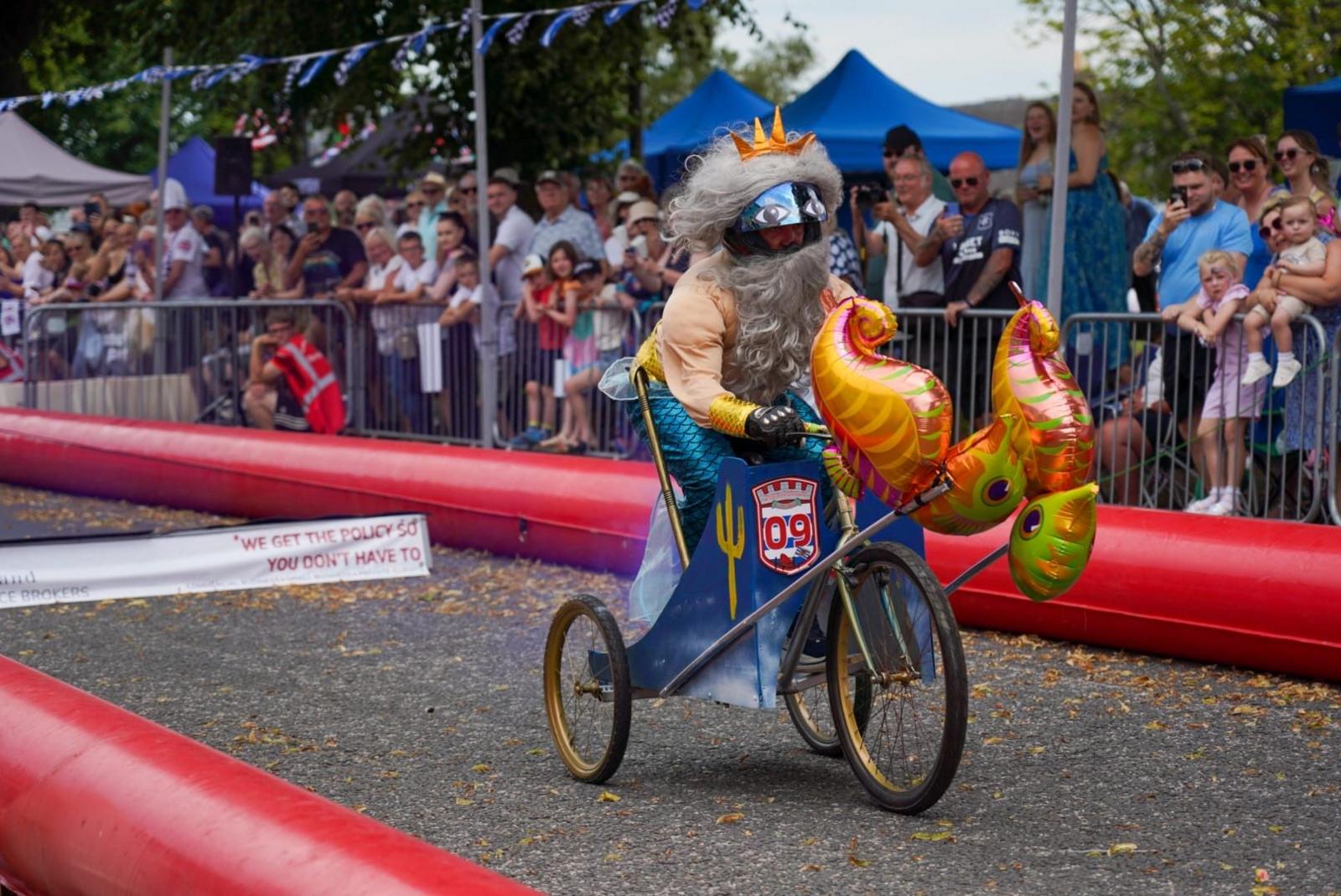 Crowds look at a merman on a tricyle. He has a large grey beard and seahorse balloon on front of the kart.