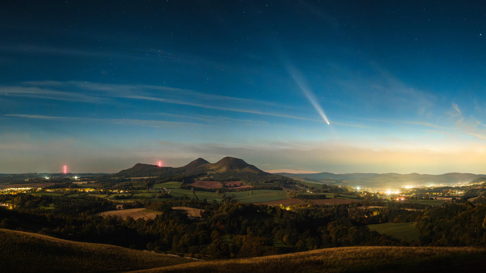 Streak of a comet in evening sky above rolling countryside and a hill with the sparkling of town lights in the distance