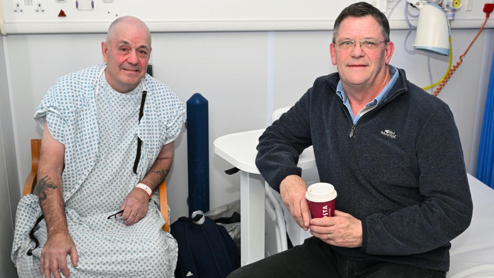 Two men sit side by side in a hospital. One is in a white plastic hospital gown after treatment. 