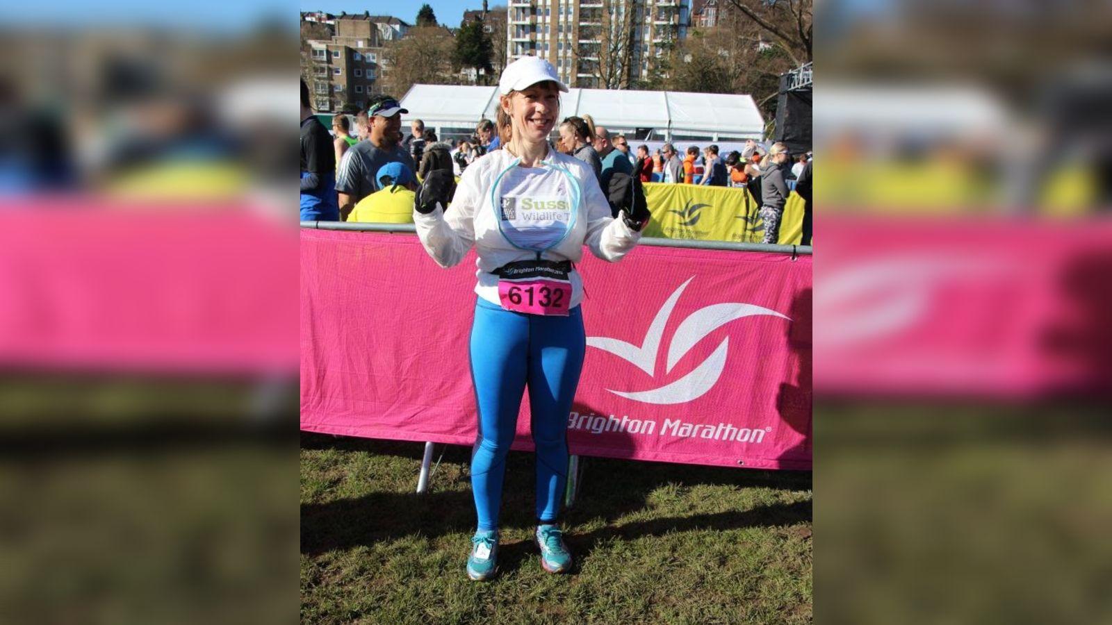 A woman poses in running gear in front of a pink banner that says 'Brighton Marathon'. She is smiling.