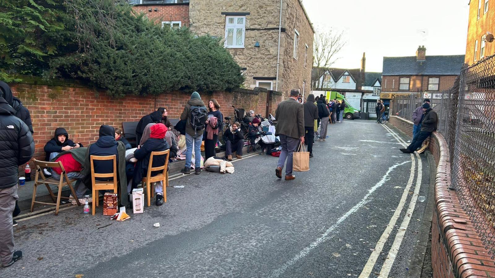 A road with a fence on one side and a brick wall on the other, with a row of people lining one side. Some are sitting on dining chairs and most are wearing hats and coats, with food and bags at their feet.