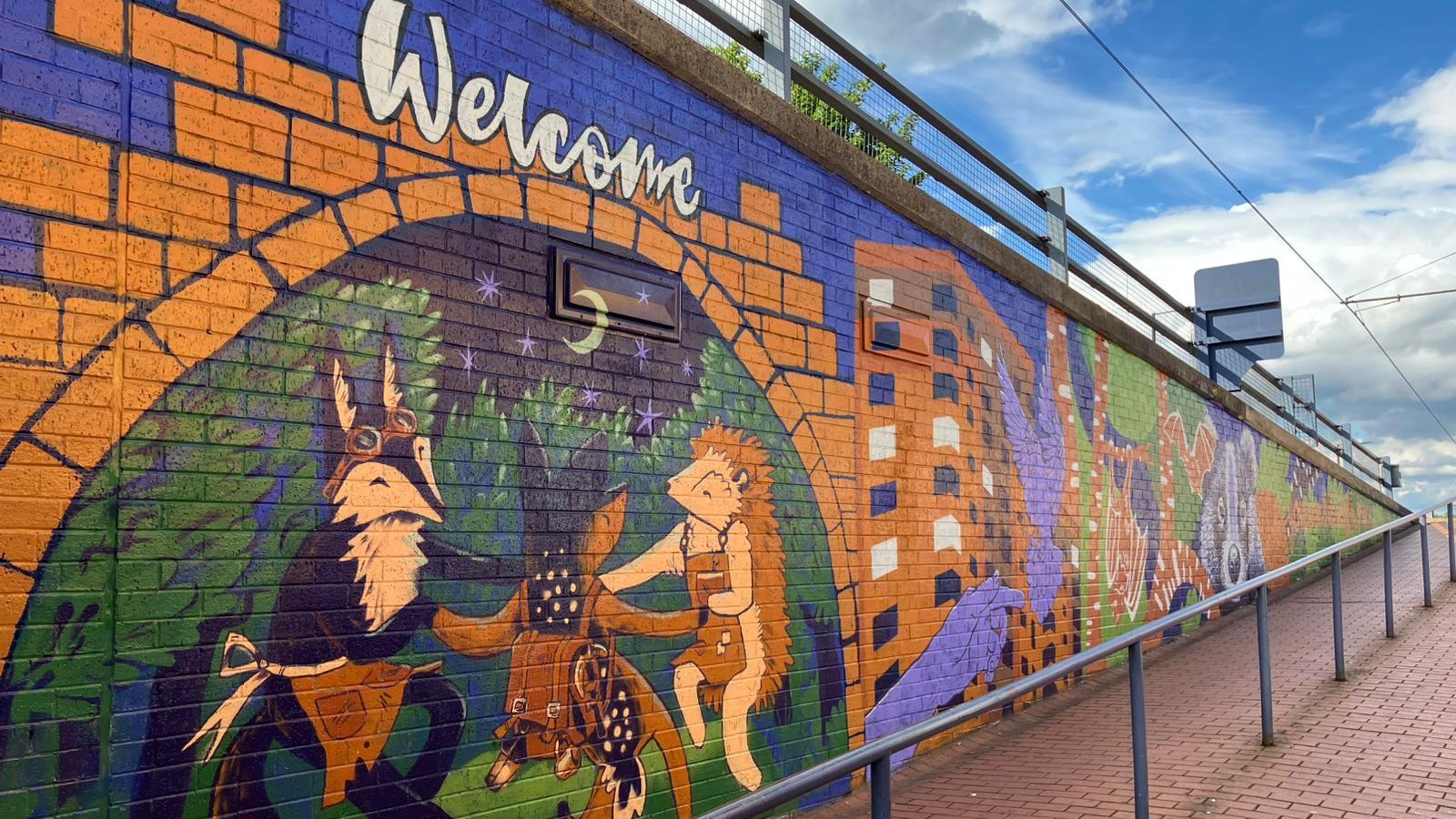 A mix of purple, green, orangey brown and dark blue makes up the cartoon-style mural. There are lots of nature themes such as a badger and hedgehog dancing, a large raccoon facing the viewer, as well as some buildings with the windows painted a mix of white and black to show lights on and off.