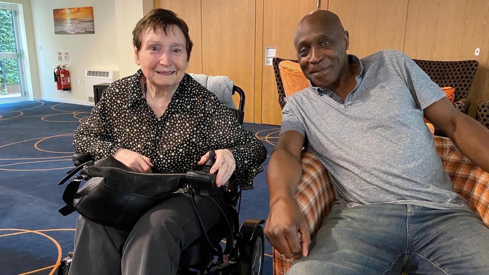 Former footballer Gary Thompson with a care home resident. She is sat in a wheelchair and has short dark hair and is wearing a dark top with spots on it. Garry sits in a tartan-print chair in a grey T-shirt and jeans