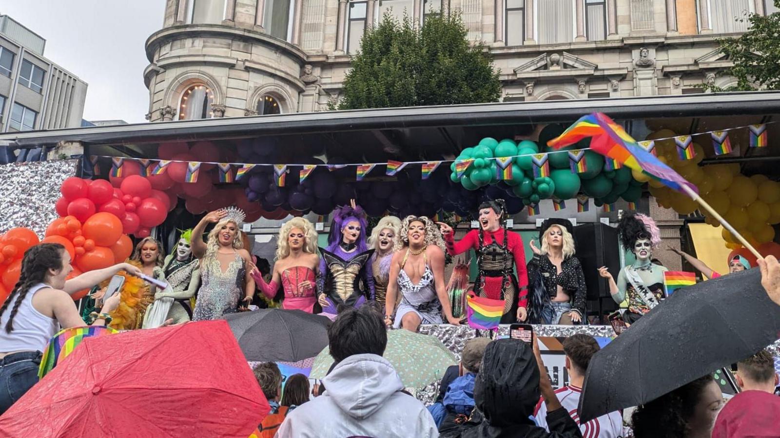 Group of drag queens on a pride float waving and pointing at the crowd