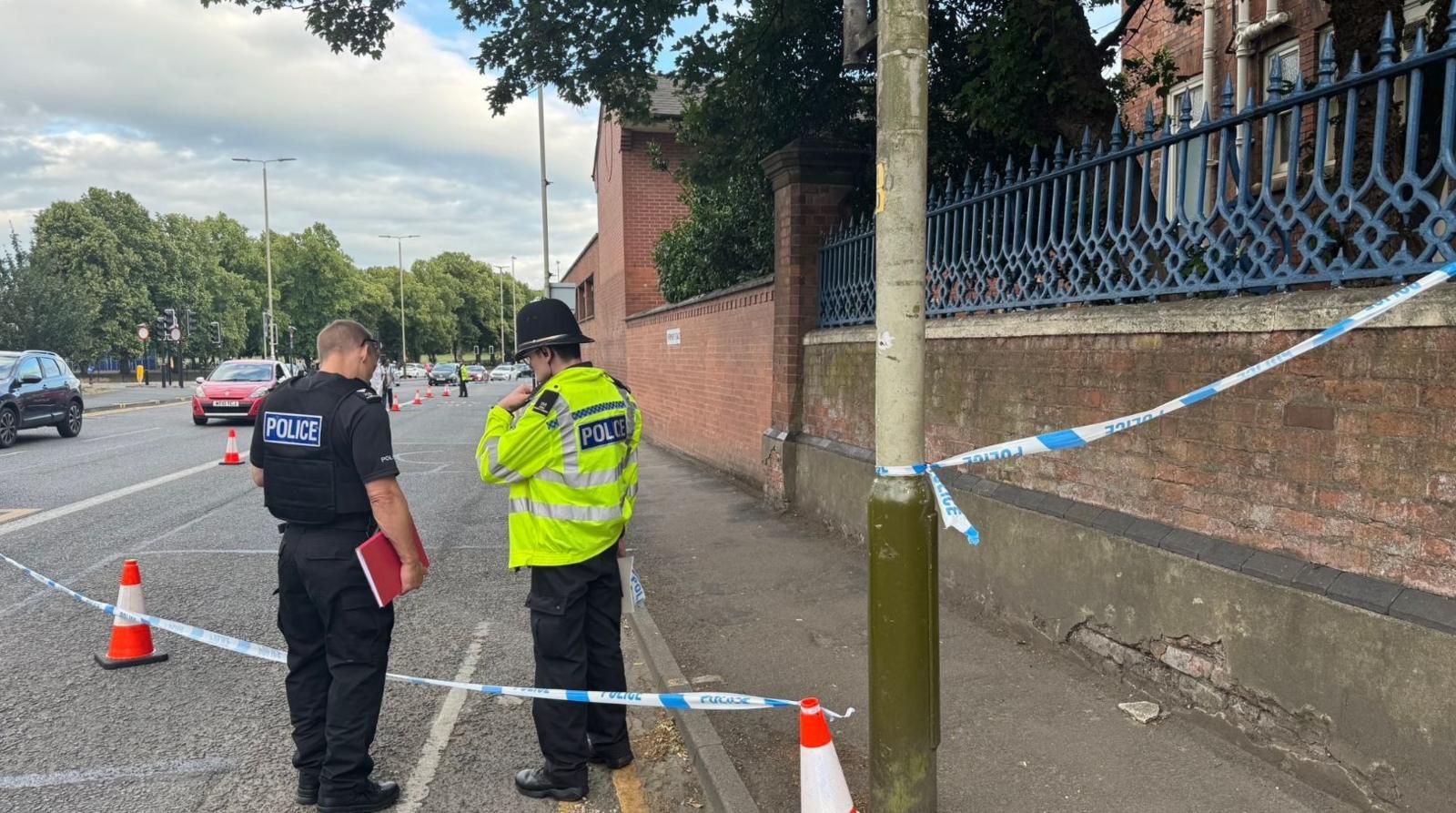 Two police officers standing by a tape cordon on the edge of a busy road
