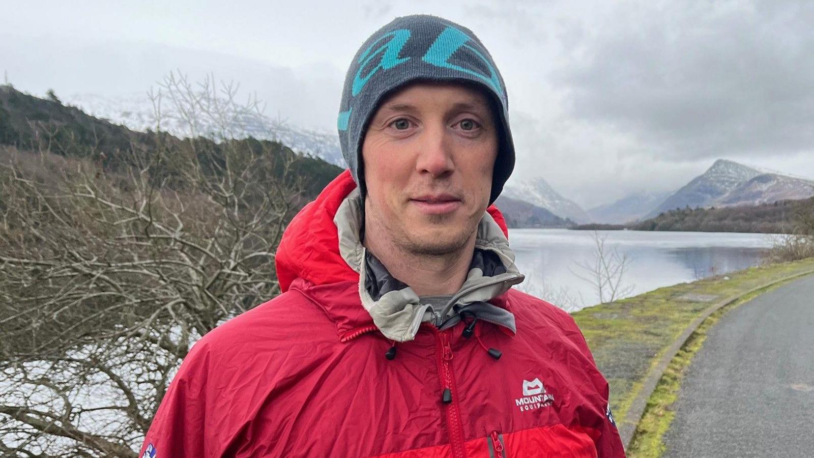 Mae Tom wedi gwisgo yn gynnes o flaen llyn a'r mynyddoedd yn y cefndir