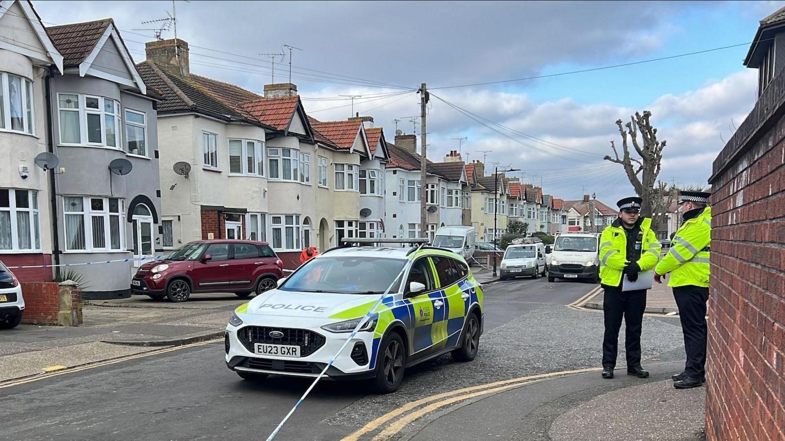 A police car by a taped off area in Southend, showing a brick wall to the right, a road, with cars parked on it, police in the distance and a row of houses to the left. There are also houses to the right. Several trees and a telegraph pole are in the road, and double yellow lines. Two police officers are to the right. 