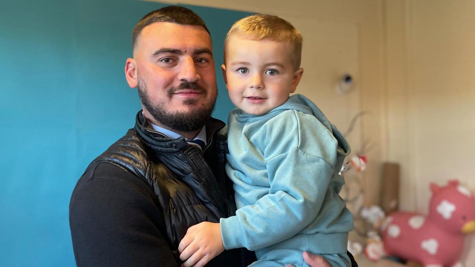 A man wearing a light blue shirt and a black jacket, with dark hair and dark facial hair, is holding a three-year-old boy. The boy is wearing a turquoise sweatshirt and has light-coloured hair.