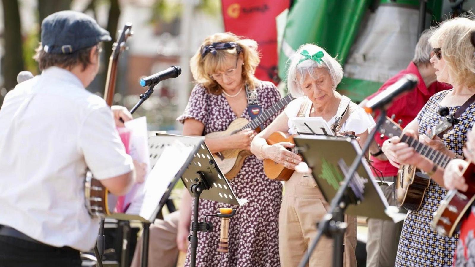 Three women and one man playing ukuleles outdoors. The man is wearing a blue cap and white shirt and is facing the women. Two of the women are wearing blue and white dresses and the other is wearing a white top and beige trousers. There are music stands and microphones in front of them.