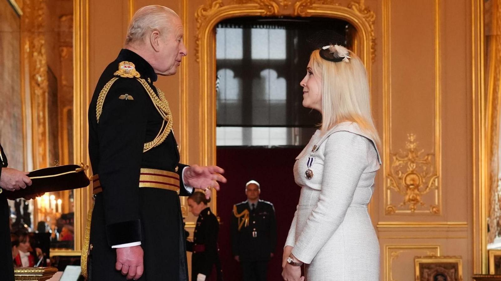 The King, dressed in a dark military uniform with gold detailing, stands facing Katherine Goodwin, who is wearing a light grey dress and a small black fascinator. They appear to be conversing during an investiture ceremony in an ornate gold-trimmed room, with a uniformed officer standing in the background.