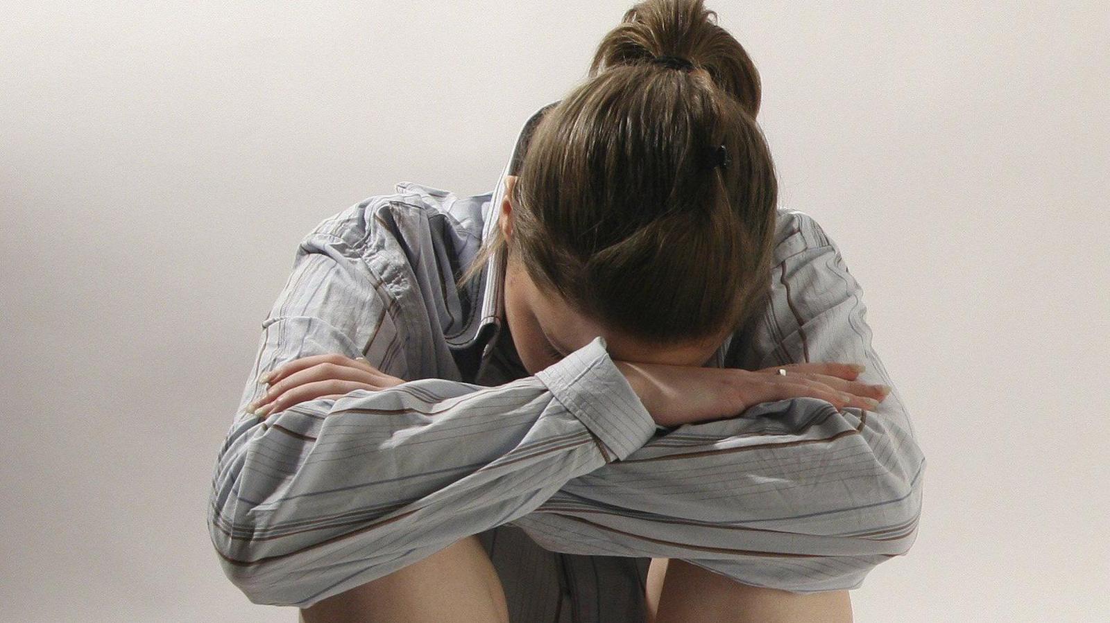 A woman sitting on the flour with her knees tucked up and her arms wrapped around them. She has her head on her arms. She is wearing a grey shirt with thin maroon stripes on it. She is wearing beige shorts. She has dark hair which is tied back in a pony tail.