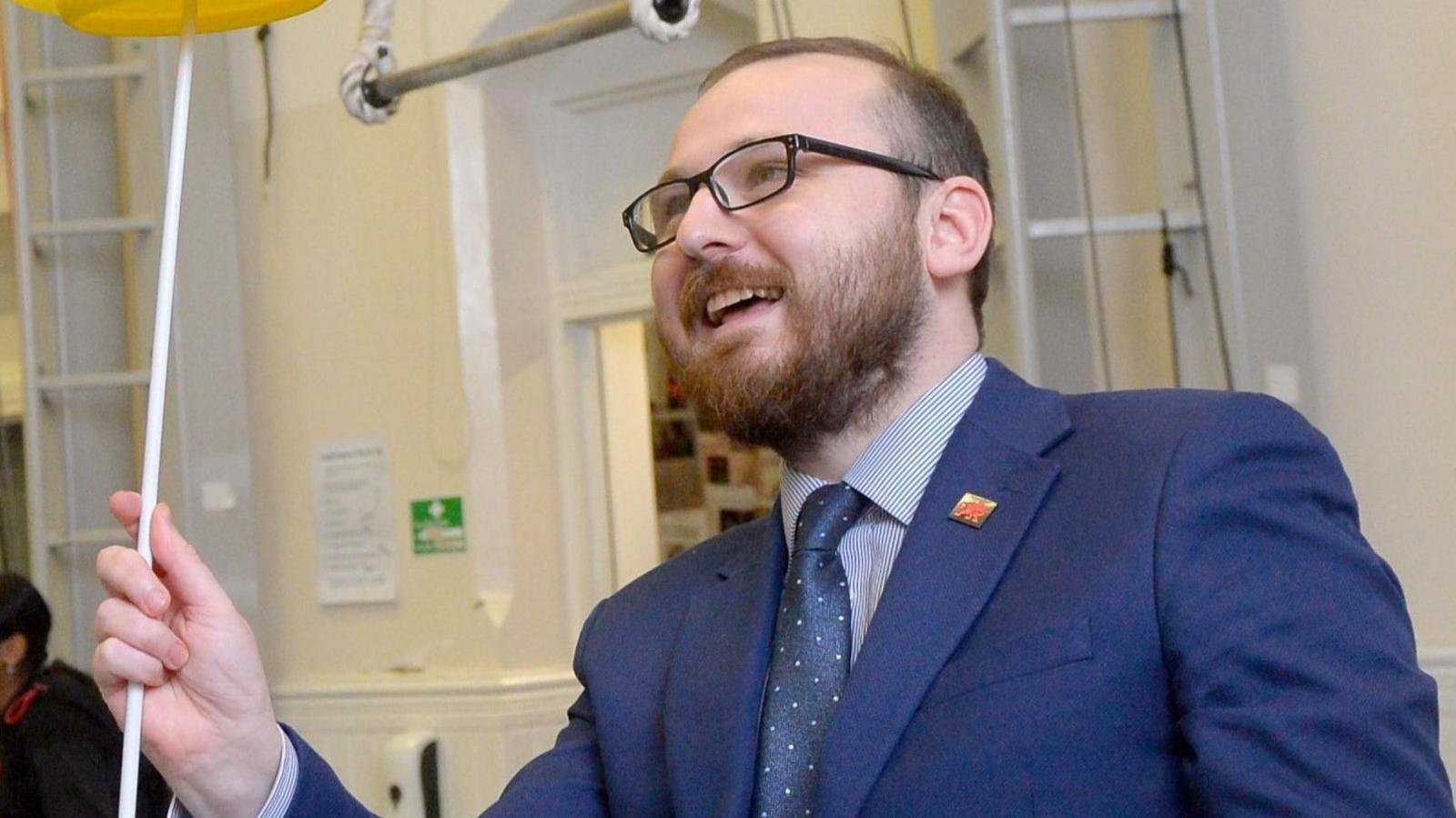 Jack Sargeant, wearing a blue suit and glasses, stood spinning a yellow plate at a circus training centre in Cardiff. He is holding a white pole, while the yellow plate is just visible at the top of the photo.