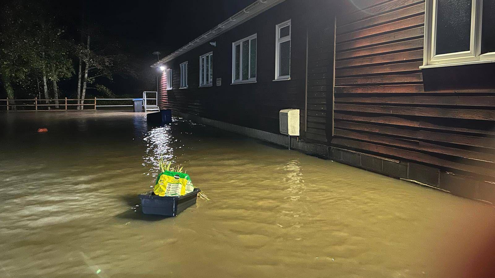 Flood water rising by a brown log building with dark skies and trees in the background.