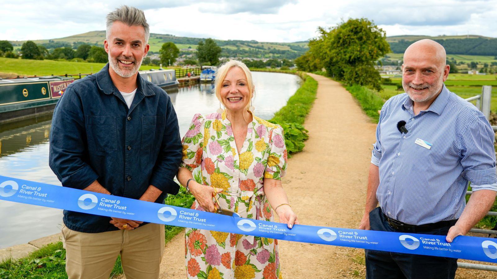 A man with grey hair and a beard who is wearing a blue long-sleeved shirt and beige chinos. He is stood next next to a woman who is wearing a colourful floral dress with blonde hair tied back and a man with a bald head who is wearing a blue and white shirt and blue trousers. They are stood in a line on a towpath next to a canal. In front of them is a blue ribbon which the woman is holding and is about to cut with a pair of scissors. 