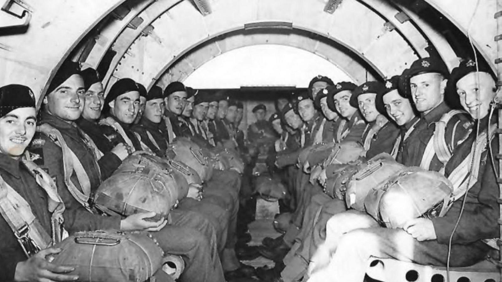 A black and white photo of around 26 RAF personnel sat on either side of the inside of a world war two plane. All the officers are wearing military uniform and holding large packs on their laps, and turn their heads to smile at the camera. The arches of the plans fuselage can be seen above their heads.