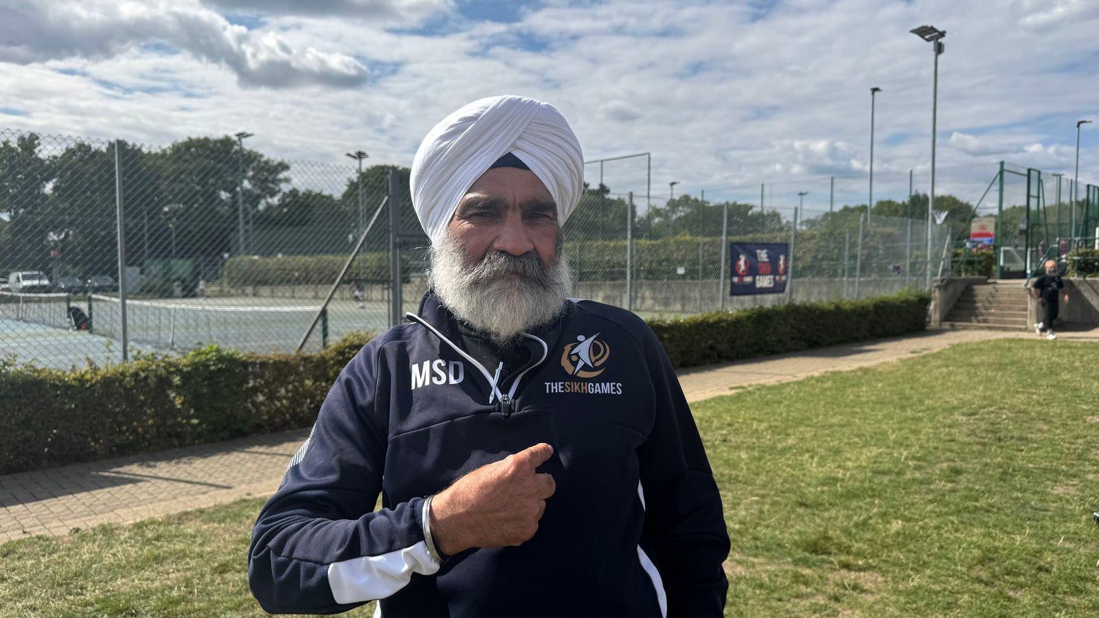 Meva has a white turban and white beard. He wears a dark blue quarter-zip sweater and is standing in front of a tennis court.