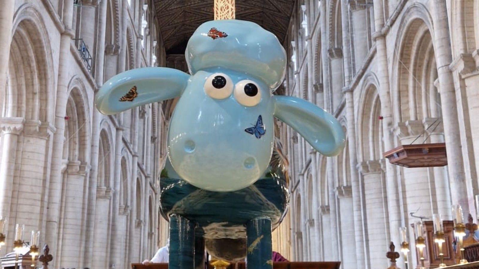 A large, light blue sheep is standing in the middle of a cathedral. Large white, ornate columns are either side of the sheep, who has butterflies on each side of his face.