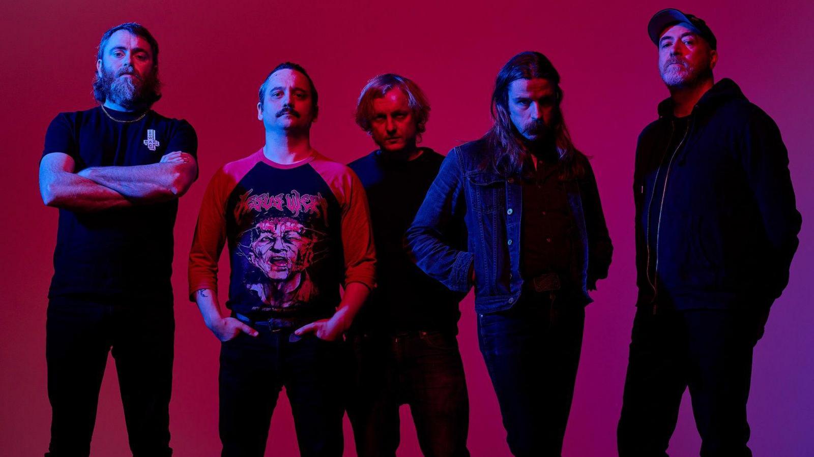 The band standing in a line in front of a red background. There is a blue light shining on them putting them largely in shadow. The man on the left has his arms folded and has a bushy beard and chin-length hair. The man next to him is wearing a red band t-shirt and has his hands in his front pocket. The man in the middle is standing slightly behind everyone else in all black. To his right the band member has long hair and has his hands in his black jacket pockets. The man on the far right is in black with his hands in his pockets and a cap. His head is tilted backwards slightly. They are all looking into the camera and all of them have straight faces.