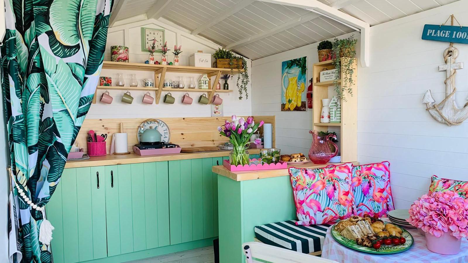 Inside the beach hut, which has green cupboards. There is various crockery on the wall above a sink and there are curtains with leaves on. To the right of the image is a table with food on top of it. A bench seat with pink pillows surrounds it.