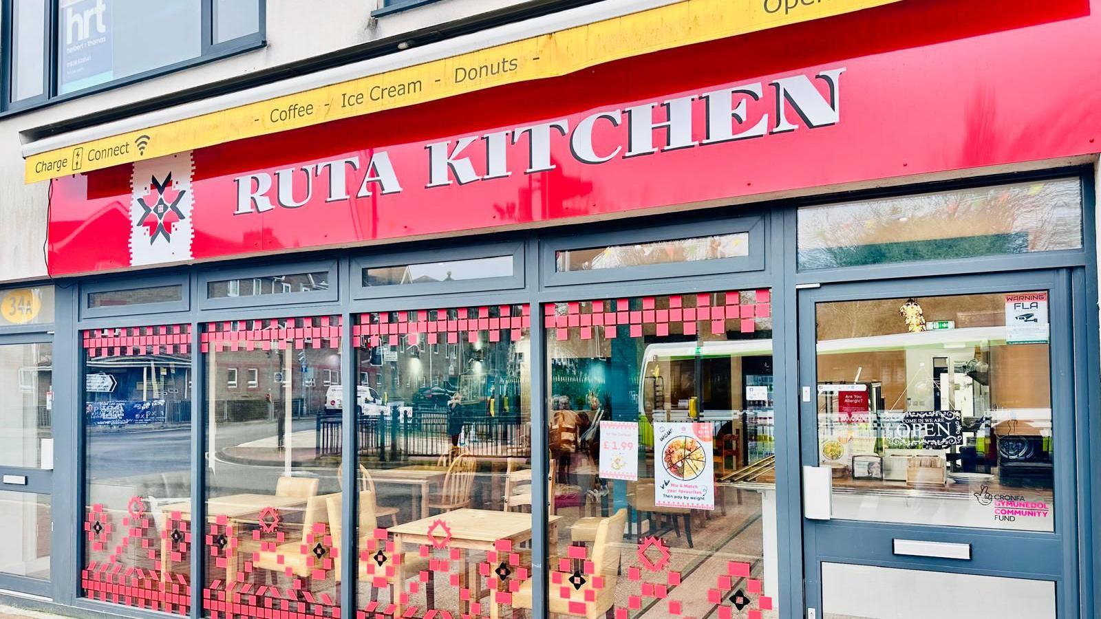 The exterior of Ruta Kitchen with a red sign and large glass windows with red decoration on them.