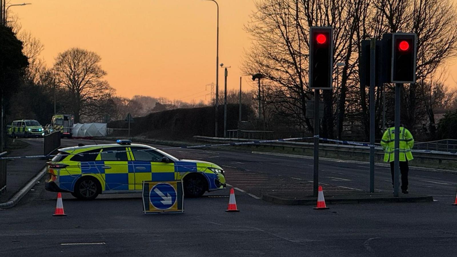 A police car is parked blocking the A5. There are also cones and police tape restricting access. The sun is rising making the sky orange. More police vehicles are parked further along the road.