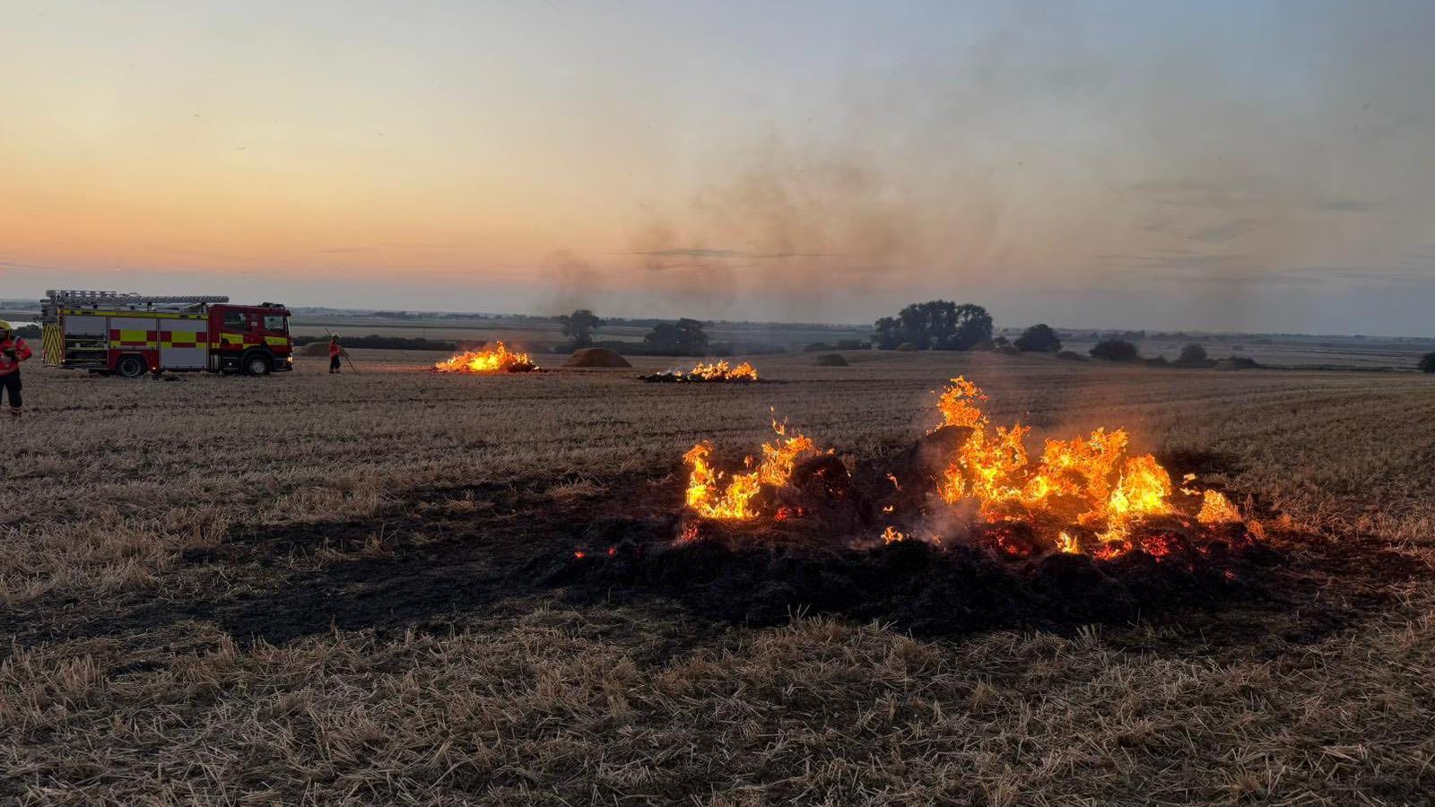 Flames and smoke billow from an area of charred grass in a field. It is almost dark. There is a fire engine parked in the background.