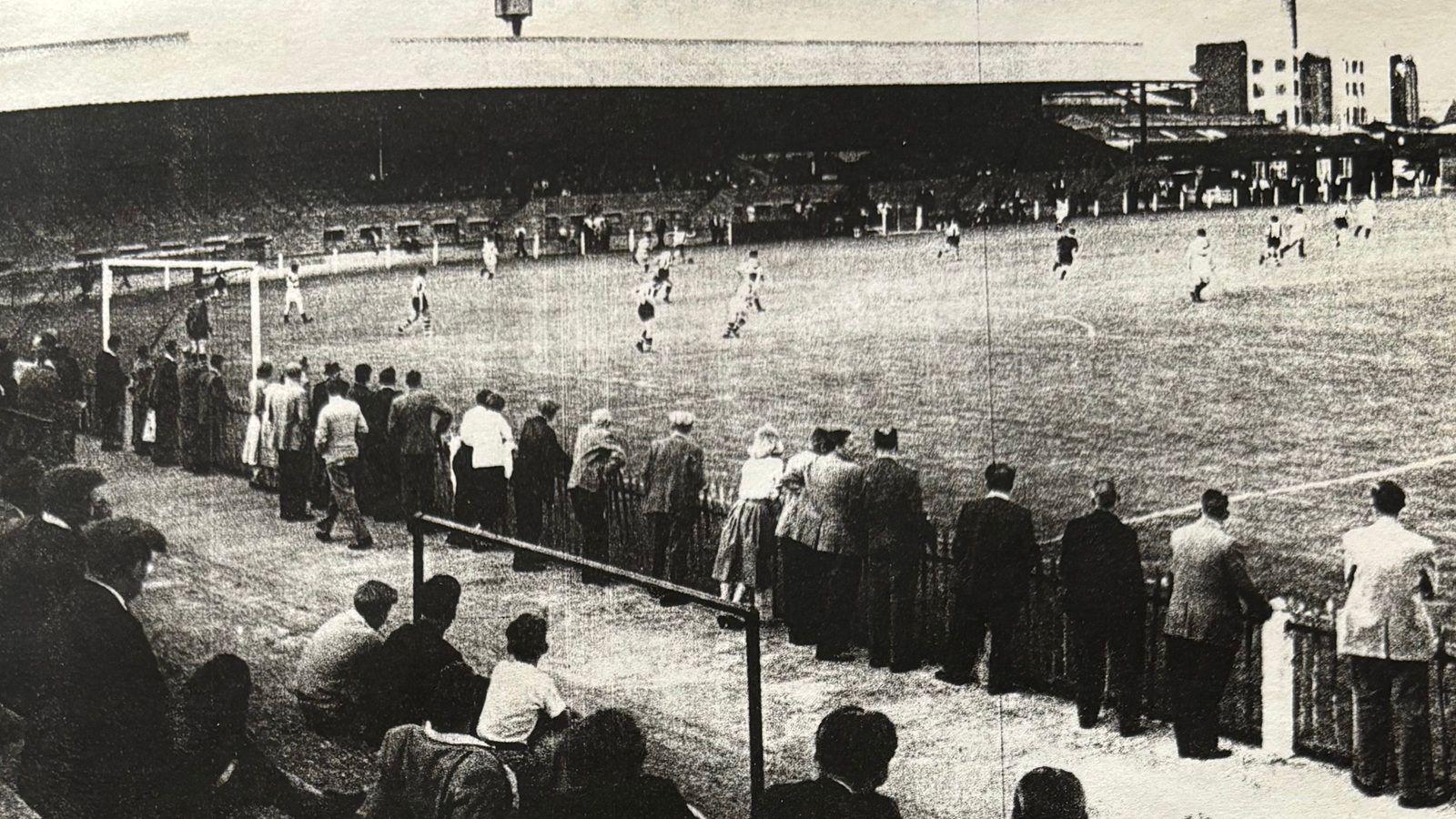 A black and white photo of a football ground, seen from one corner looking across to approximately halfway along the pitch. Along the far side runs a covered terrace, with players on the pitch running away from the photographer's angle. In the foreground of the image is some uncovered terracing, with people sitting and standing on it.