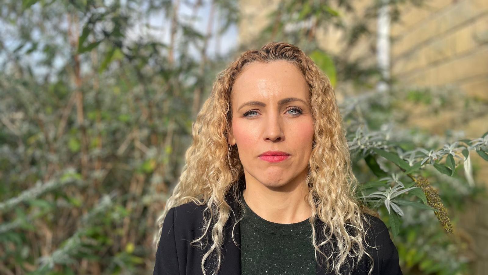 Labour MP Katie White, a woman with blonde curly hair, who is wearing a dark sparkly top and black suit jacket. She is stood against the backdrop of some trees.