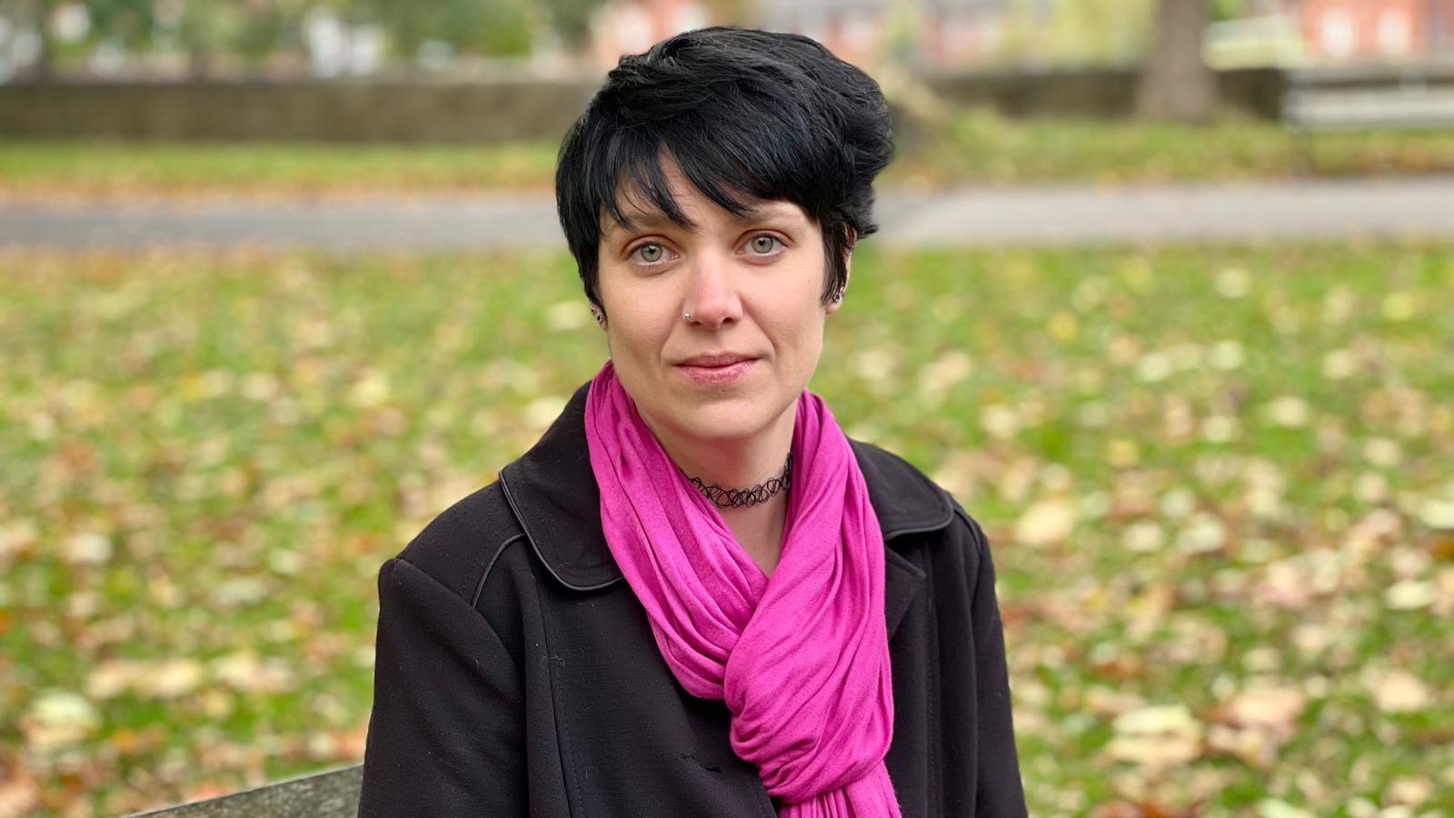 Rebekah looks at the camera sitting on a wooden bench in a park. She has short black hair and is wearing a dark pink scarf, a black choker and a black jacket