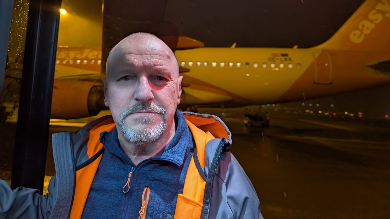 A man wearing a navy fleece and orange-lined raincoat looks at the camera as he stands on an airport bus, in front of an easyjet plane on a runway.