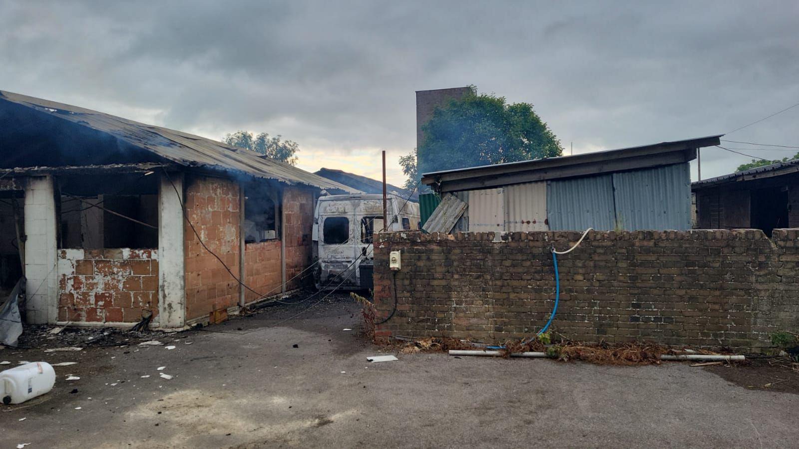 A burnt out-minivan next to what appears to be a fire-damaged workshop. Smoke can be seen rising from the structure. 