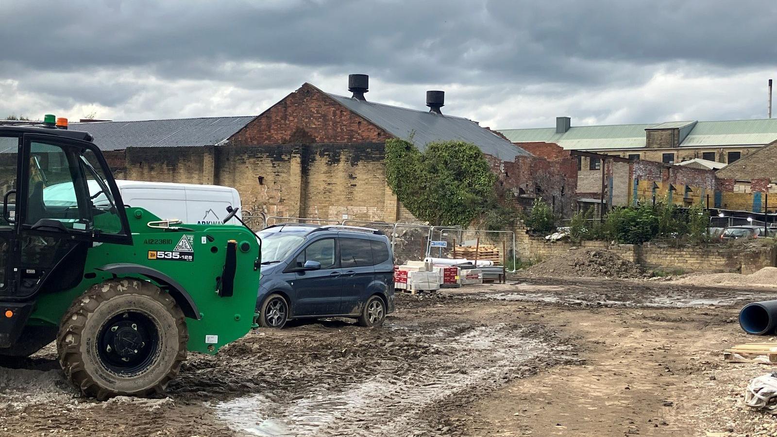 A plot of apparent wasteland. A tractor and a 4x4 sit nearby. Some ramshackle old buildings are in the background.