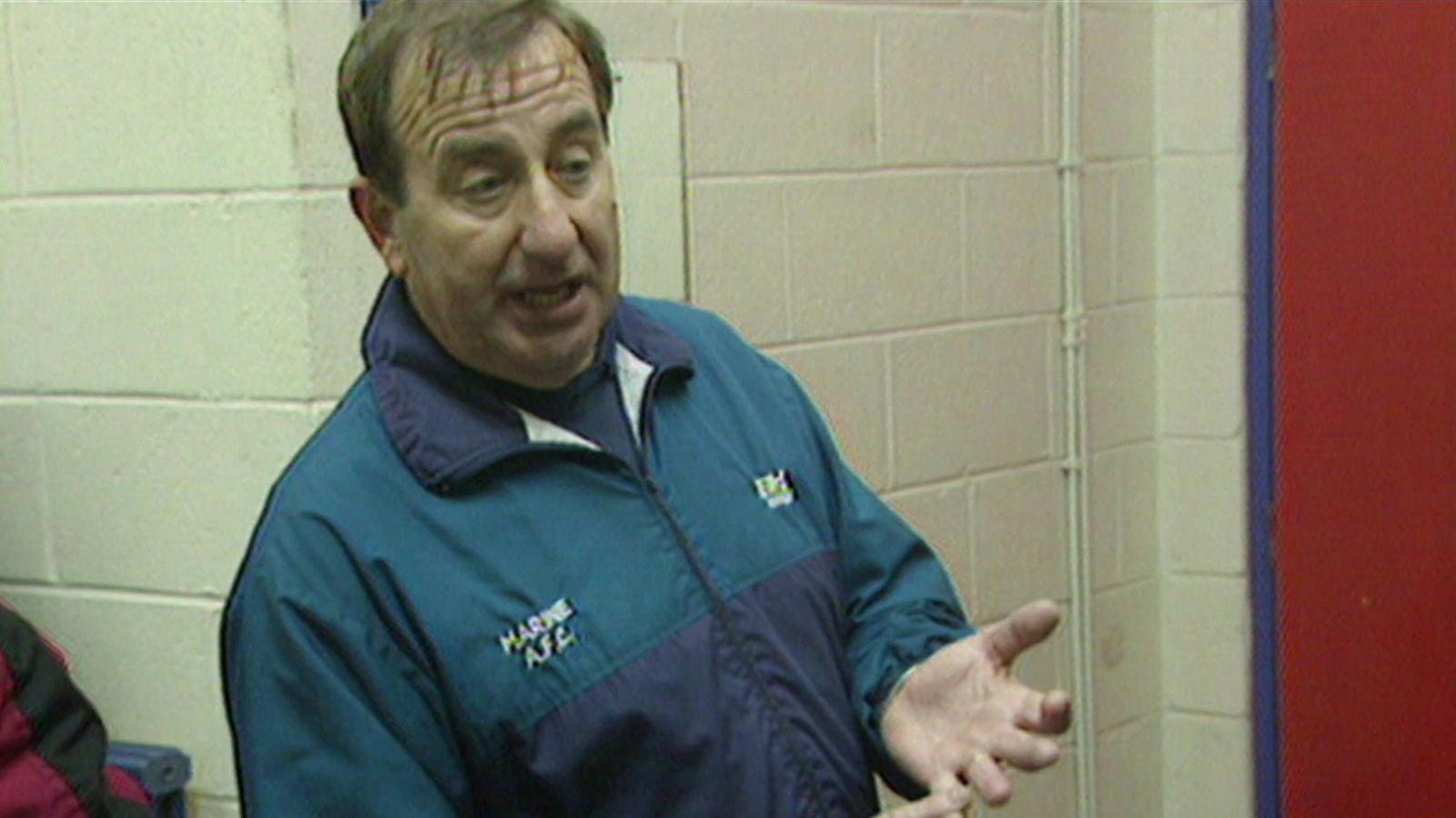 Roly Howard, wearing a turquoise, holds his hands out while standing in a footballers' dressing room. He has short brown hair. 