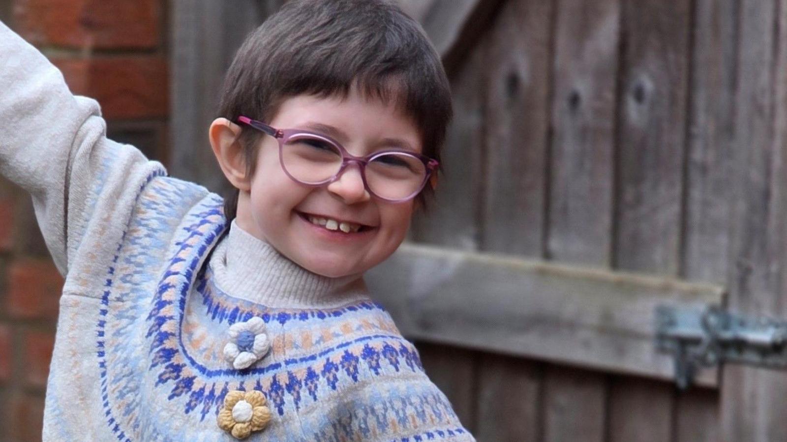 A young girl with short dark hair, purple-framed glasses and a grey patterned jumper, smiling, with one arm raised, in front of a brick wall with a wooden gate.