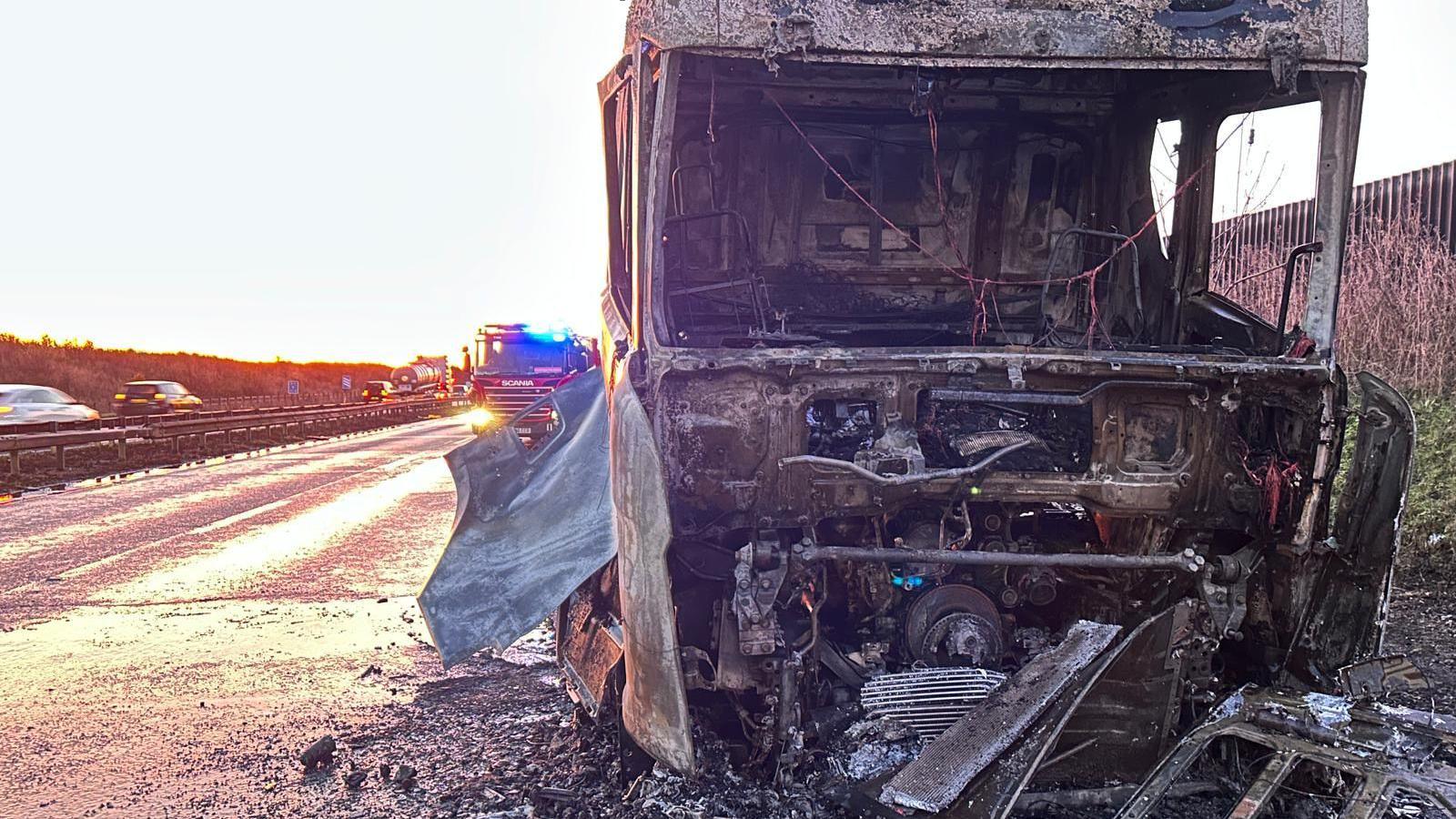 A lorry on the side of the M11 has been burnt and melted. Directly inside the metal framing of the seats can be seen and the metal work around the vehicle has been charred. 