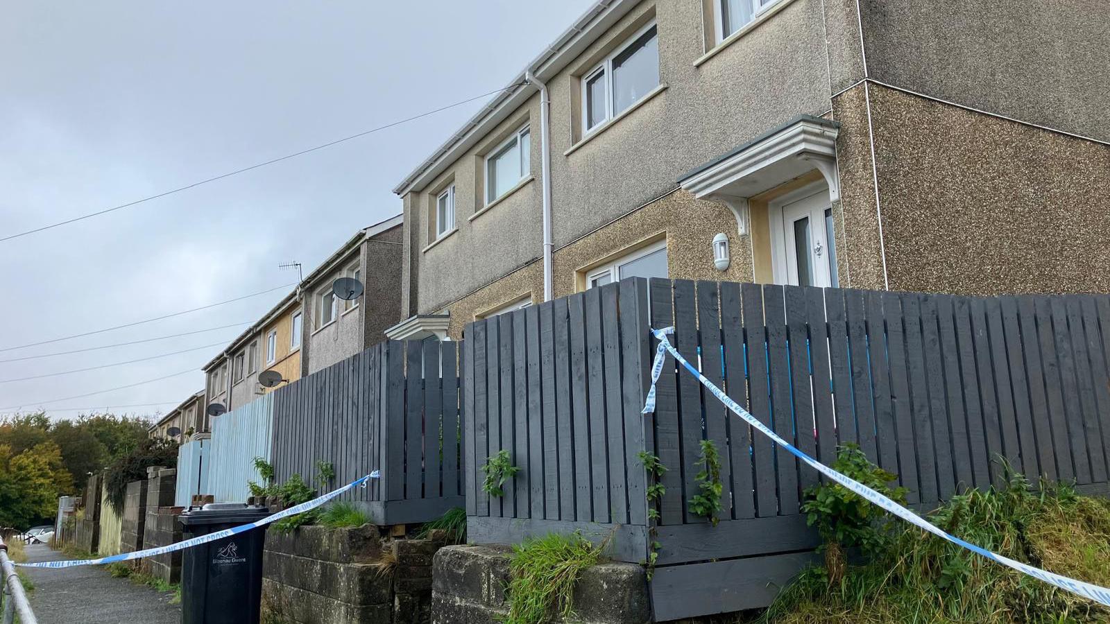 Semi-detached pebble dashed terraced house with wooden fence surrounding the front garden and steps down to the pavement. Police tape seals off the walkway alongside the house and part of the pavement in front.