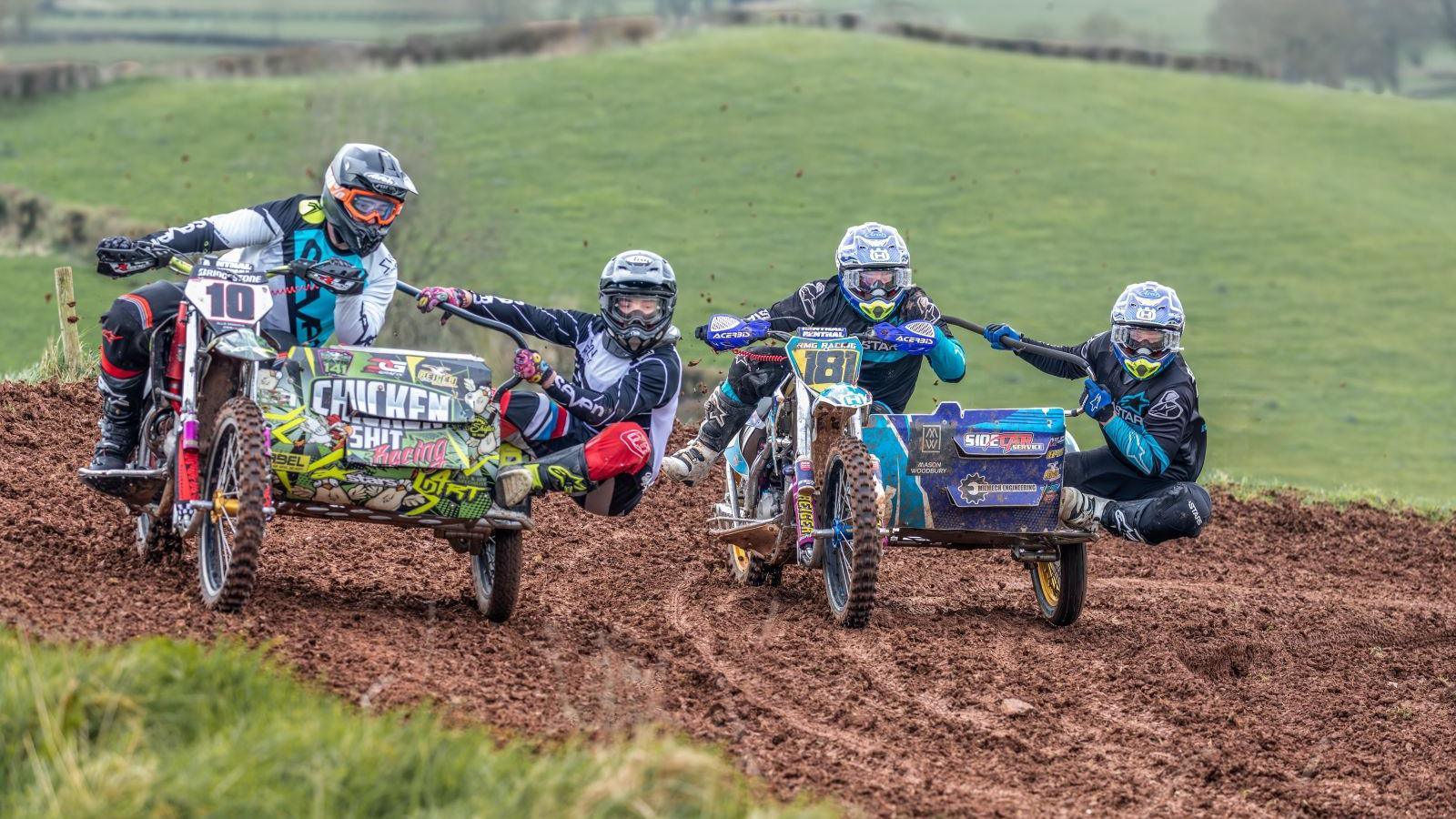 Four people wearing biker outfits are riding two bikes with boxes. One person each is riding the bike, while another hangs of the side of the box. They are going across a dirt path over fields.