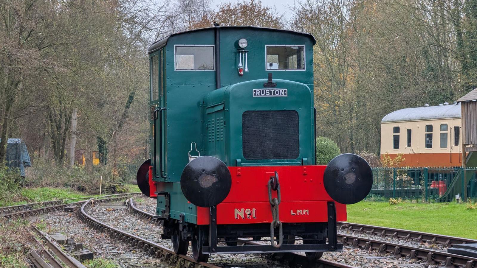 Mr Rose's Sir William McAlpine Ruston 48 moves down a track. Trees surround the small section of tracks while other passenger train carriages can be seen in the background. 