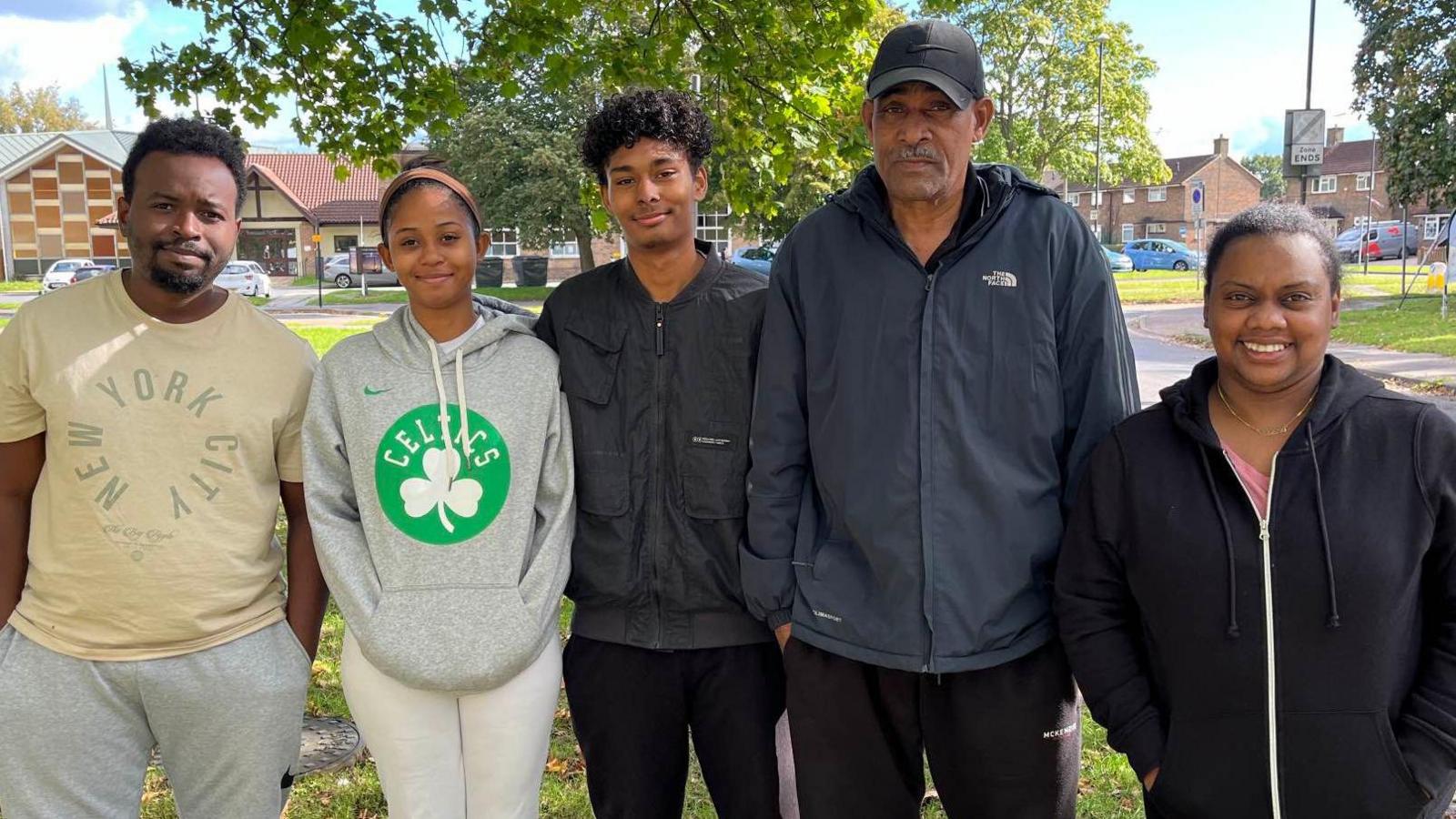 Five people in a line. Left to right there is a man in a yellow shirt and grey joggers, a woman in a grey hoodie with a Boston Celtics logo on it and yellow trousers, a man in a black zip-up jacket and black trousers, an older man in a navy North Face windbreaker and black joggers, and a woman with a black zip-up hoodie