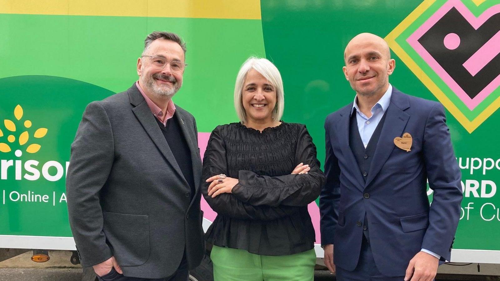 A line of three people from the left, a man wearing a grey suit jacket and black V-neck jumper and pink shirt, a woman with silver hair, black blouse and green trousers and a man with a bald head wearing a blue suit jacket and light blue shirt under a dark blue cardigan. 