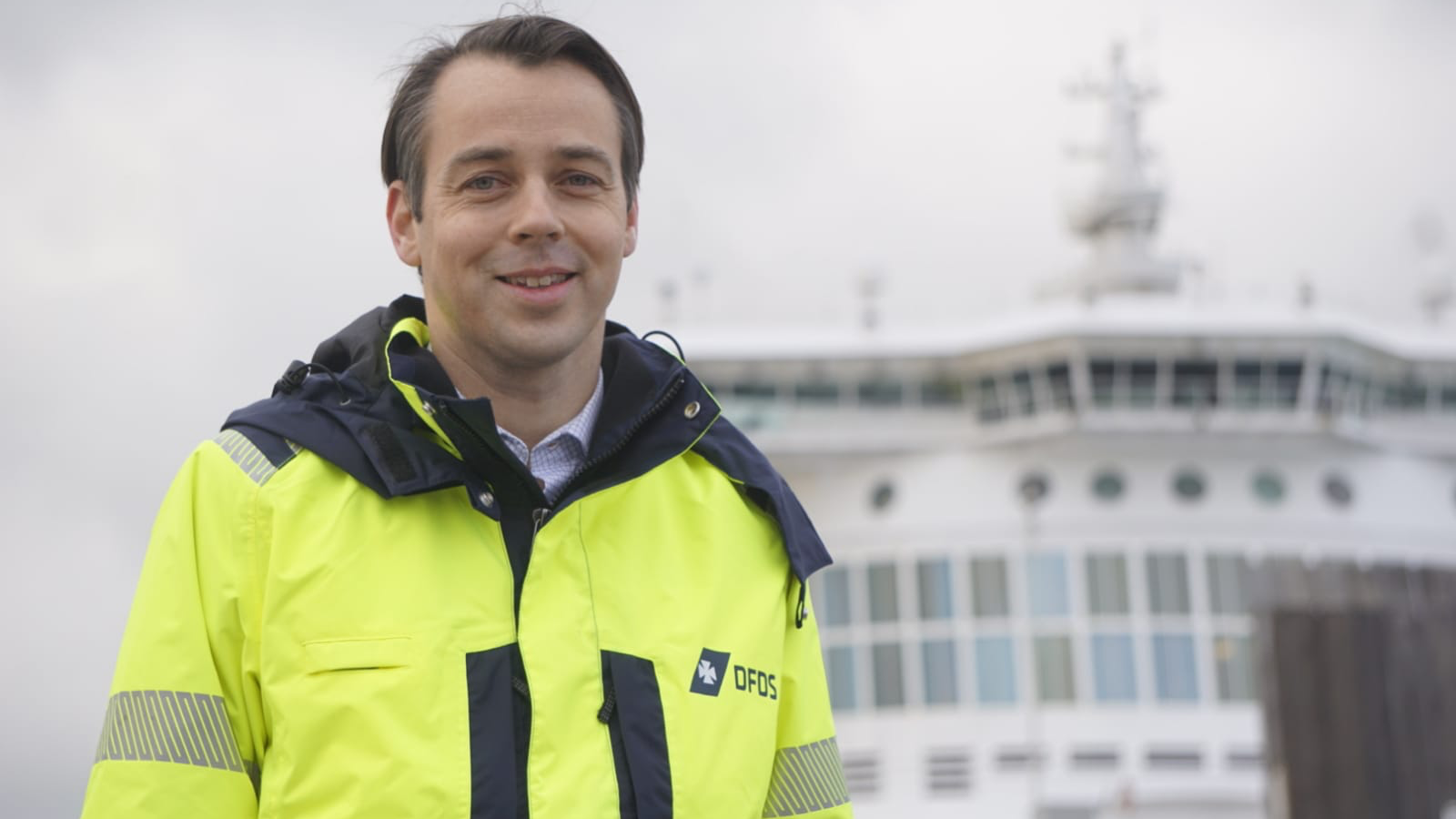 Filip Werne Hermann smiles at the camera and is standing on the deck of a large boat. He is wearing a fluorescent yellow anorak with DFDS printed on the breast pocket.