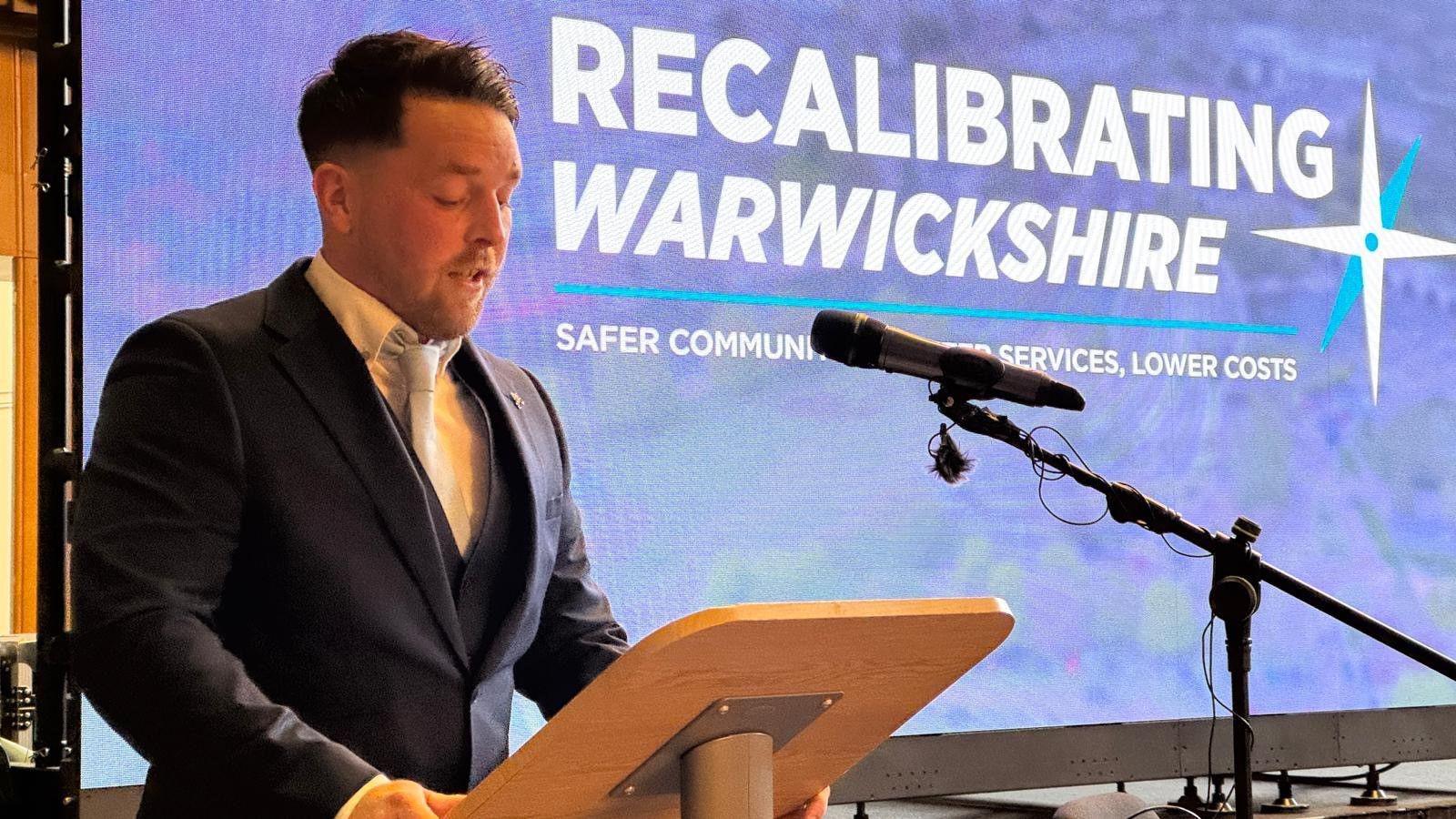 A man A man in a suit standing at a lectern with a microphone delivering a speech. He is in a navy suit and silver tie. There is large screen behind him with a purple background the reads "Recalibrating Warwickshire".