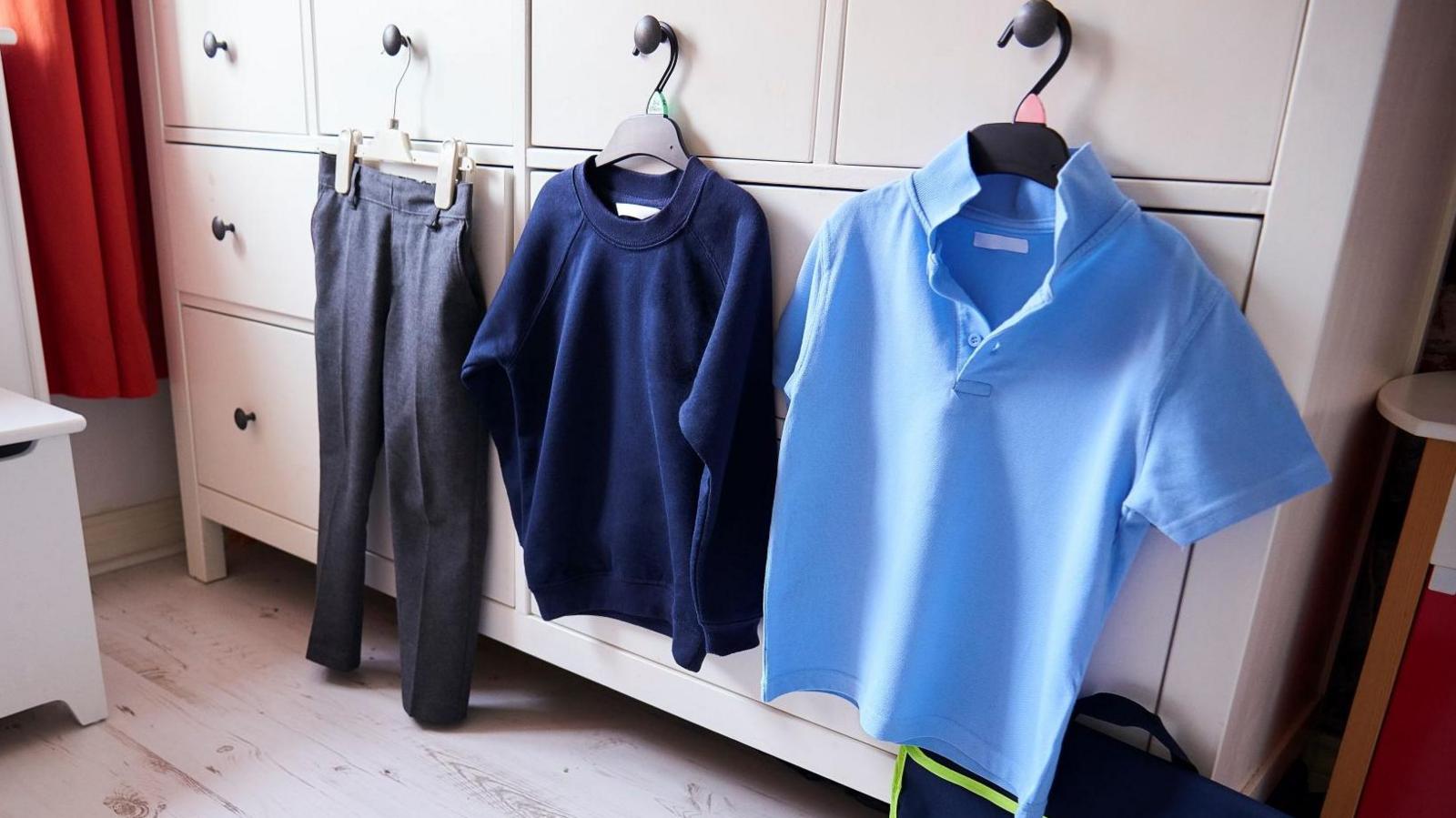 A child's light blue polo shirt, a navy crew neck jumper and a pair of grey trousers hang on three separate hangers on a white chest of drawers.