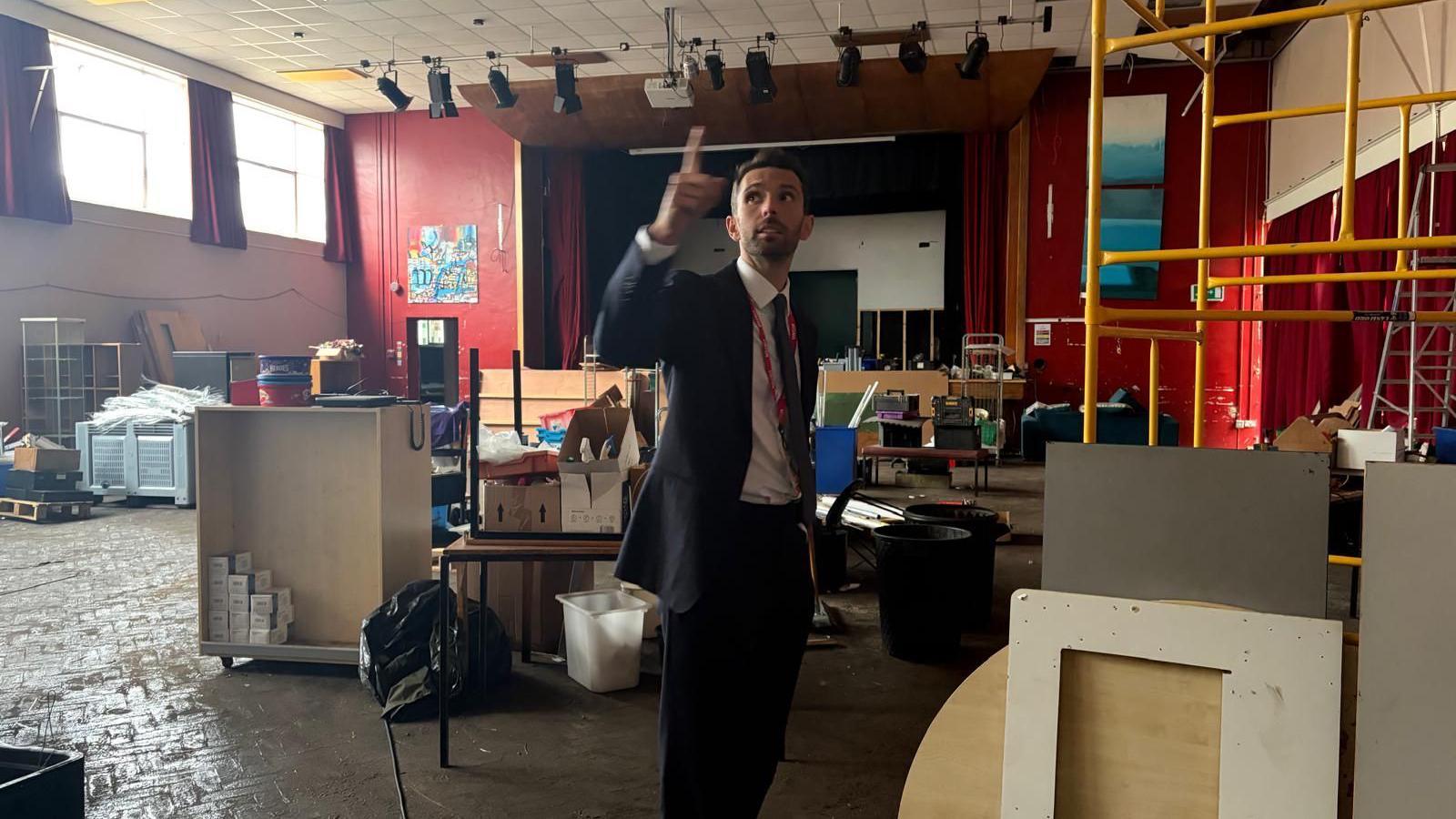 Minister Josh MacAlister stands in the hall of Myton School, where there are lots of cabinets and equipment scattered around whilst work is done on the school to remove RAAC. He is standing wearing a suit and pointing up at the ceiling.