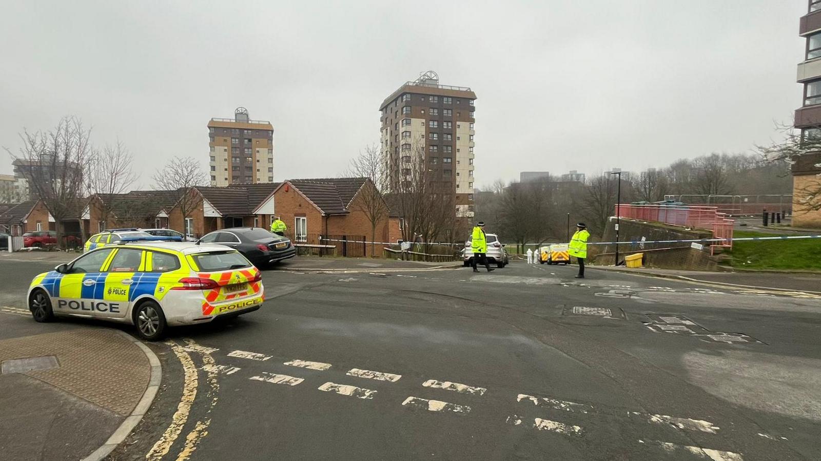 Three police officers guard a cordon that stretches from one side of the road to the other. A police car is parked on the left. Three skyscrapers can be seen in the background.