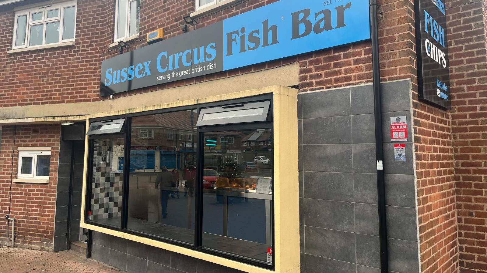 The outside of Sussex Circus Fish Bar. It has a blue and black sign above the door and window